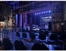 Innenraum eines kleinen Konzertsaals mit einer Bühne, die mit Instrumenten und Beleuchtung ausgestattet ist. Im Vordergrund stehen Tische und Stuhlreihen, während im Hintergrund eine Leinwand mit der Aufschrift „SCHWIPSA – Das Jubiläumskonzert, 23. März 2024“ zu sehen ist.