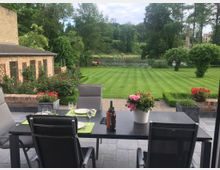 Terrasse mit schwarzem Esstisch und grauen Stühlen, gedeckt für eine Mahlzeit, mit Blumen als Dekoration. Im Hintergrund ein gepflegter Rasen, ein kleiner Teich, umgeben von Bäumen, und ein Backsteingebäude an der linken Seite.