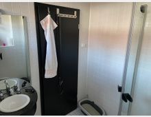 Ein Badezimmer mit weißen Wandfliesen, einem schwarzen Waschtisch mit zwei Waschbecken und einem Spiegel. Im Vordergrund hängt ein weißes Handtuch an einer dunklen Tür, rechts befindet sich eine Katzentoilette und eine Duschkabine mit Glastüren.