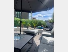 Moderne Dachterrasse mit Holzboden, mehreren grauen Polstermöbeln und einem großen Sonnenschirm. Im Hintergrund befinden sich Pflanzen, ein kleiner Pool und ein Haus mit großen Glasfenstern.