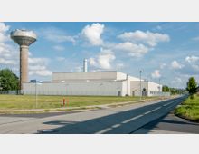 Industriegebiet mit einer modernen Lager- oder Fabrikhalle aus Beton, einem hohen Wasserturm im Hintergrund und einer angrenzenden Straße. Das Gelände ist von einer Grünfläche und einem Metallzaun umgeben, bei klarem Himmel und vereinzelten Wolken.