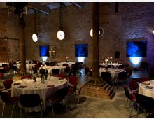 Ein rustikaler Veranstaltungsraum mit unverputzten Backsteinwänden, sichtbaren Holzsäulen und moderner Beleuchtung. Mehrere runde Tische sind mit weißen Tischdecken, Kerzenständern und Geschirr für ein Abendessen eingedeckt, umgeben von roten Stühlen.