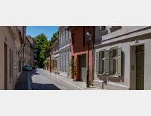 Schmale gepflasterte Gasse mit historischen Wohnhäusern in unterschiedlichen Farben, darunter Weiß, Beige und Rot, mit Fensterläden und Laternen. Im Hintergrund sind Bäume zu sehen, teils unter blauem Himmel.