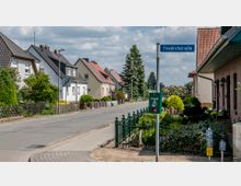 Eine ruhige Wohnstraße mit Reihen von Ein- und Mehrfamilienhäusern, gepflegten Vorgärten und einem Gehweg. Ein Straßenschild mit der Aufschrift „Friedrichstraße” ist sichtbar, daneben ein Mülleimer für Hundeabfälle und eine Geschwindigkeitsbegrenzung auf 30 km/h.