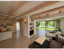 Offener Wohnraum in einem modernen Haus mit sichtbaren Holzbalken an der Decke, einer Einbauküche links, einem Wohnzimmer mit Sofas rechts und einer großen Glasfront, die auf eine Terrasse mit Blick auf einen grünen Garten und einen See führt.