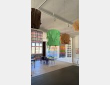Ein heller, moderner Innenraum mit hohen Decken, dekoriert mit großen, hängenden Lampenschirmen aus bunten Materialien wie grünem Stoff und Stroh. Im Hintergrund stehen Tische und Stühle, während halbtransparente Vorhänge mit farbigen Streifen den Raum optisch trennen.