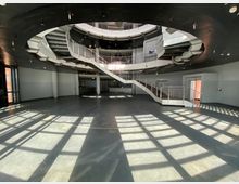 Großes, modernes Atrium mit einem kreisförmigen Grundriss, einer geschwungenen Metalltreppe in der Mitte und mehreren Ebenen. Tageslicht fällt durch Fenster ein und wirft Schattenmuster auf den glatten Fußboden.