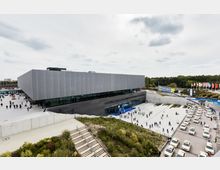 Moderne Konferenz- oder Veranstaltungshalle mit grauer, minimalistischer Architektur und großen Glasflächen im Erdgeschoss. Im Außenbereich befinden sich ein gepflasterter Platz mit Besuchern, Treppen, Grünflächen sowie eine Straße mit parkenden Taxis; im Hintergrund wehen Nationalflaggen.