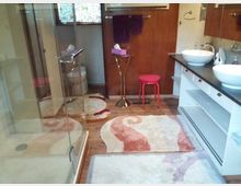 Modernes Badezimmer mit Holzboden und Glasdusche. Es verfügt über zwei weiße Aufsatzwaschbecken auf einer dunklen Arbeitsplatte, bunte Teppiche, eine Toilette und einen pinken Hocker nahe einer Holzwand.