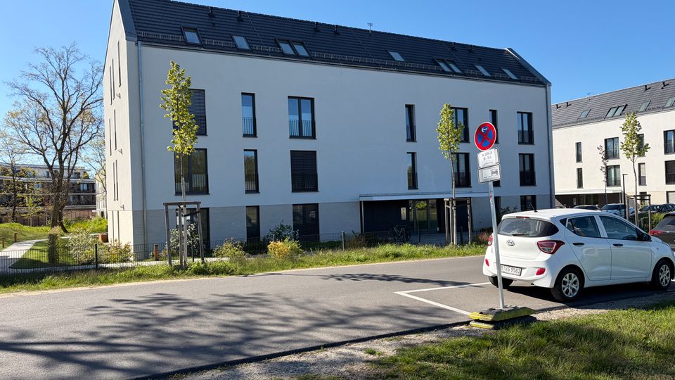 Modernes, dreigeschossiges Wohngebäude mit weißer Fassade, dunklem Satteldach und schmalen Fenstern entlang einer asphaltierten Straße. Im Vordergrund stehen ein Verkehrszeichen, ein geparktes Auto sowie junge Straßenbäume und Grünflächen; daneben sind weitere ähnliche Wohnblöcke zu sehen.