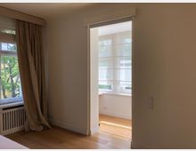 Ein heller Innenraum mit weißen Wänden und einem Holzboden, der teilweise einen Durchgang zu einem angrenzenden Raum mit großen Fenstern zeigt. Links befindet sich ein Fenster mit beigen Vorhängen, durch das Bäume draußen sichtbar sind.