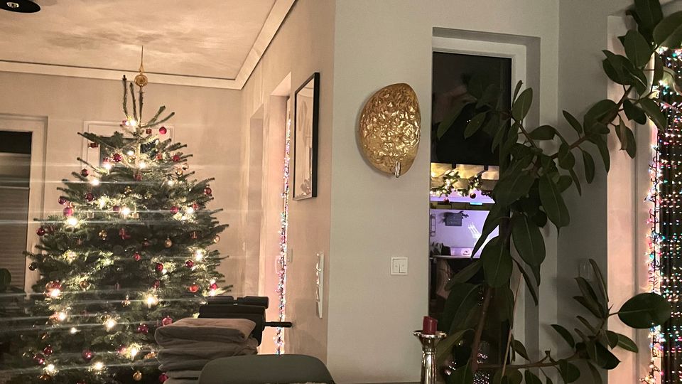 Ein Wohnbereich mit festlich geschmücktem Weihnachtsbaum, der mit Lichtern und Kugeln dekoriert ist. Im Vordergrund steht ein Esstisch mit Platzsets, Kerzenständer und einigen Papieren, im Hintergrund sind eine Topfpflanze und bunte Lichterketten sichtbar.