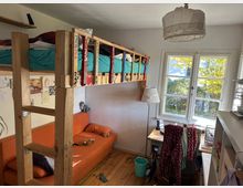 Kleines, helles Kinderzimmer mit einem Hochbett aus Holz, einer orangenen Couch darunter und einem Schreibtisch vor einem Fenster mit Blick auf Bäume und Herbstlaub. An der Wand hängen Zeichnungen, und ein Regal sowie eine Stehlampe ergänzen die Einrichtung.
