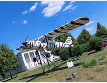Ein kleines Flugzeug mit Zebrastreifen-Muster steht als Ausstellung auf einer Metallkonstruktion in einem Freigelände. Im Hintergrund sind ein weißes, modernes Gebäude, Bäume und ein blauer Himmel zu sehen.