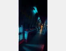 Dunkler Flur mit futuristischem Design, beleuchtet von blauem und rotem Licht; links Wände mit gitterartigen Strukturen, rechts Konsolen mit leuchtenden roten Anzeigen und einem beleuchteten Gang im Hintergrund.