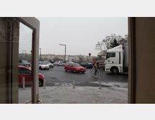 Blick durch eine geöffnete Tür auf eine städtische Straße mit viel Verkehr, darunter Autos, ein Lastwagen und ein Fahrradfahrer. Im Hintergrund sind mehrstöckige Wohnhäuser und kahle Bäume sichtbar, bei bedecktem Himmel.