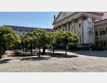 Platz mit einer gepflasterten Fläche und Reihen kleiner Bäume, die Schatten spenden. Im Hintergrund steht ein klassizistisches Gebäude mit Säulen und Dreiecksgiebel, daneben ein Gebäude mit roten Ziegeln und großen Fenstern.