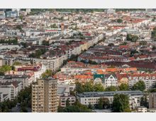 Luftaufnahme einer Stadt mit dicht bebautem Wohngebiet, geprägt von roten Ziegeldächern, hellen Fassaden und vereinzelten modernen Gebäuden. Im Hintergrund ist eine Kirche mit spitzem Turm sowie grüne Parkflächen zu erkennen.