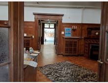 Innenraum eines Gebäudes mit Holzvertäfelung und einem Kamin auf der rechten Seite. Im Hintergrund führt eine Tür zu einem helleren Bereich mit Pinnwänden und Pflanzen. Ein durchsichtiger Glastisch mit Schreibutensilien sowie ein Schild mit der Aufschrift „Bitte nicht rauchen“ sind ebenfalls sichtbar.