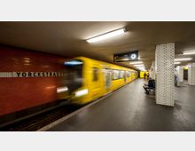 U-Bahn-Station Yorckstraße in Berlin mit gelben Zügen, roten Kacheln an der Wand und weißen Fliesen an den Säulen. Eine elektronische Anzeigetafel mit Uhr sowie sitzende und stehende Personen sind ebenfalls sichtbar.