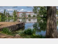 Blick auf einen ruhigen See mit einem Betonsteg im Vordergrund, umgeben von grüner Vegetation und einem Baum rechts im Bild. Im Hintergrund stehen mehrstöckige Wohngebäude mit roten Dächern und Balkons, teilweise von Bäumen verdeckt.