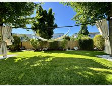 Grüne Gartenlandschaft mit gepflegtem Rasen, umgeben von Büschen und Bäumen. In der Mitte hängt eine weiße Hängematte zwischen zwei Bäumen, von denen cremefarbene Vorhänge seitlich befestigt sind; im Hintergrund ist ein Holzzaun, Häuser und blauer Himmel sichtbar.