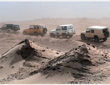 Eine Reihe von Geländewagen fährt durch eine sandige Wüstenlandschaft mit sichtbaren kleinen Dünen im Vordergrund. Der Himmel wirkt staubig, und die Umgebung ist karg und ohne Vegetation.