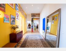 Ein Flur mit gelben Wänden, geschmückt mit zahlreichen bunten Bildern. Ein großer Spiegel steht an der rechten Wand, ein Teppich liegt zentral auf dem Holzboden. Bücherregale und eine Türöffnung mit Blick auf ein Klavier und weitere Räume befinden sich im Hintergrund.