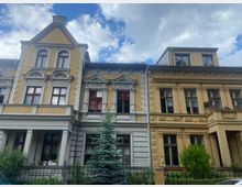 Zwei nebeneinander stehende Altbauhäuser mit gelblichen Fassaden, roten Fenstern und dekorativen Details wie Säulen und Gesimsen. Vor den Häusern befinden sich ein kleiner Garten, ein Metallzaun und ein Nadelbaum, darüber bewölkter Himmel.