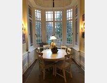 Helle Sitznische in einem Wohnraum mit großen Sprossenfenstern, durch die ein Garten mit Bäumen sichtbar ist. Im Zentrum steht ein runder Esstisch mit sechs Stühlen, einer Hängelampe und einer dekorativen Vase; die Wände sind cremefarben mit Wandlampen und Bildern.