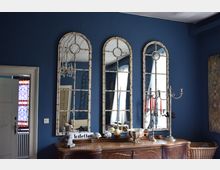 Innenraum mit dunkelblauer Wand, drei hohen, bogenförmigen Spiegeln mit weißen Rahmen und einem antiken hölzernen Sideboard im Vordergrund. Auf dem Möbelstück stehen dekorative Objekte, ein Kerzenhalter und ein Schild mit der Aufschrift "Toiletten"; im Hintergrund sind ein Kronleuchter und ein Raum mit Fenstergardinen sichtbar.