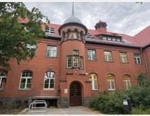 Backstein-Gebäude mit roten Dachziegeln und einer runden Erker-Struktur, die von einem kleinen spitzen Turm gekrönt wird. Vor dem Gebäude befinden sich Bäume, Sträucher und ein gepflasterter Weg.