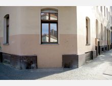 Ecke eines beigefarbenen Wohngebäudes mit braunen Fliesen im unteren Bereich, mehreren rechteckigen Fenstern und einem gepflasterten Gehweg. In einem der Fenster spiegelt sich ein benachbartes Gebäude mit roten Dachziegeln.