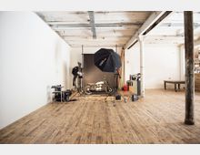 Ein Fotostudio in einem offenen Raum mit Holzboden und weiß gestrichenen Wänden. Im Mittelpunkt steht eine Motorradaufnahmeszene, umgeben von Studiobeleuchtung, Stativen und einem schwarzen Hintergrund.