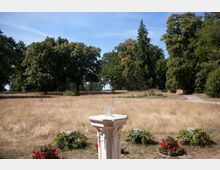 Stein-Sonnenuhr in einem Park mit trockenem, goldgelbem Gras, umgeben von Bäumen und blühenden Pflanzen in Blumenkübeln im Vordergrund, unter einem klaren blauen Himmel. Ein schmaler, geschwungener Weg führt durch die Grünfläche.