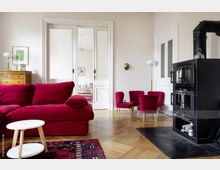 Wohnzimmer mit klassischem Interieur: Rote Samtmöbel, ein weißer Beistelltisch und ein bunter Teppich prägen den Raum; ein schwarzer Kaminofen steht rechts, während eine Flügeltür mit Glasscheiben in einen angrenzenden Raum führt. Der Raum hat helle Wände, Holzboden und vereinzelt dekorative Elemente wie Lampen und Bilder.