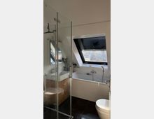 Modernes Badezimmer mit Dachschräge, einem großen Dachfenster mit Rollladen, einer Badewanne, einer verglasten Dusche, einem weißen Waschbecken mit Holzunterschrank und dunklen Fliesen auf dem Boden.