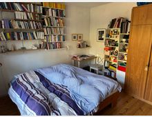 Schlafzimmer mit Holzfußboden, einem Bett mit gestreifter Bettdecke im Vordergrund und mehreren Bücherregalen an der Wand. Rechts im Raum befinden sich ein Schreibtisch mit Stuhl, ein Regal mit Ordnern und Gegenständen sowie ein großer Holzschrank.