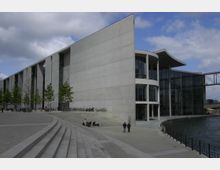 Modernes Regierungsgebäude mit einer Fassade aus Beton und Glas, großen, geschwungenen Fenstern und einem angrenzenden Fluss. Vor dem Gebäude befinden sich breite Treppen, ein gepflasterter Platz und einige Fahrradständer.