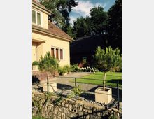 Gelbes Einfamilienhaus mit rotem Ziegeldach und einer kleinen Eingangstreppe aus Backstein, umgeben von einem gepflegten Garten mit Rasenfläche, Pflanzkübeln mit kleinen Nadelbäumen und einer niedrigen Steinumrandung. Im Hintergrund sind Bäume und ein weiteres dunkles Gebäude zu sehen.