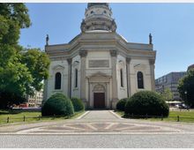 Historische Kirche mit einer Kuppel, flankiert von Säulen und schmuckvollen Fenstern, umgeben von gepflegten Sträuchern und geprägten Pflastersteinen. Links und rechts befinden sich Bäume und moderne Gebäude im Hintergrund.