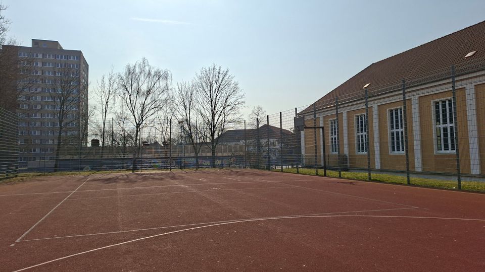 Eingezäunter Sportplatz mit rotem Kunststoffbelag und aufgemalten Spielfeldlinien, umgeben von kahlen Bäumen. Rechts steht ein langes, gelbes Gebäude mit Satteldach und hohen Fenstern, links ist ein mehrstöckiges Hochhaus im Hintergrund zu sehen.