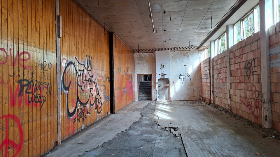 Verlassener, langer Innenraum einer Industriehalle mit hoher Betondecke, teilweise zerstörtem Boden und Graffiti an einer Holzvertäfelung sowie an Backsteinwänden. Rechts fällt Tageslicht durch eine Reihe hoher Fenster; am Ende führt eine Türöffnung zu einer Treppe.