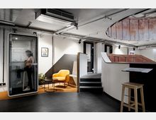 Moderner Bürobereich mit einer schallisolierten Telefonkabine auf der linken Seite, daneben ein gelber Sessel und ein runder Beistelltisch. Im Hintergrund befinden sich Stufen, ein erhöhter Arbeitsbereich mit rötlicher Metallverkleidung und sichtbare Deckenkonstruktionen mit Rohren und Spotlights.