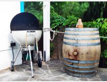 Ein Grill mit offenem Deckel steht auf einer gepflasterten Fläche neben einem Holzfass, auf dem eine kleine Tonflasche steht. Im Hintergrund sind grüne Kletterpflanzen sowie ein Holzzaun zu sehen.