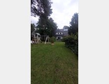 Ein Einfamilienhaus mit Satteldach und Dachfenstern steht am Ende eines gepflegten Gartens mit Rasenfläche. Links steht ein Holzspielplatz und ringsherum sind hohe Bäume und Sträucher zu sehen.