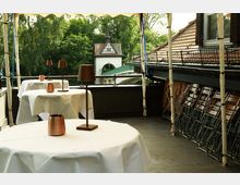 Überdachte Restaurantterrasse mit runden Tischen in weißen Tischdecken und schlanken Tischlampen; seitlich stehen zusammengeklappte Stühle an einer Hauswand mit Ziegeldach. Im Hintergrund sind Bäume sowie ein kleiner Turm- und Torbogenbau neben einer Brücke zu sehen.