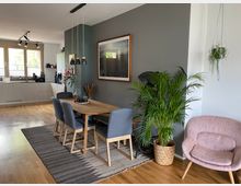 Wohnzimmer mit Essbereich und Küche in offenem Grundriss. Ein Holztisch mit grauen Stühlen und darüber hängende Pendelleuchten steht auf einem Teppich; daneben Pflanzen im Topf, eine rosa Sessel und eine graue Wand mit dekorativem Bild. Im Hintergrund eine helle Küche mit großen Fenstern.