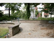 Kleiner gepflasterter Platz mit geschwungenen niedrigen Backsteinmauern, umgeben von Bäumen und Grünflächen. Im Hintergrund sind mehrstöckige Gebäude und eine Backsteinkirche mit einem auffälligen Turm sichtbar.