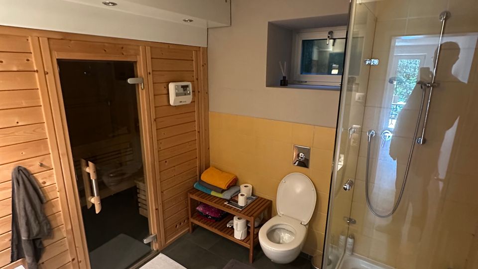 Modernes Badezimmer mit dunklen Bodenfliesen, Glasduschkabine und Toilette; links befindet sich eine holzverkleidete Sauna mit Glastür. Ein Waschbeckenunterschrank, ein kleines Wandfenster und ein Ablageregal mit Handtüchern ergänzen den Raum.